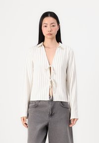 Hvit langermet topp med tynne vertikale striper, krage med knapper og frontlås med knute. Uformell passform, strukturert stoff og avslappet design.