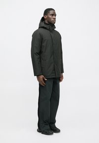 Giacca nera imbottita con zip frontale, cappuccio e tasche laterali, indossata con pantaloni scuri e scarpe nere, in piedi di profilo su uno sfondo neutro.