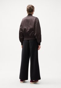 Brown bomber jacket with ribbed cuffs and hem, paired with wide black pants. Model wears burgundy sneakers, with a smooth texture backdrop.
