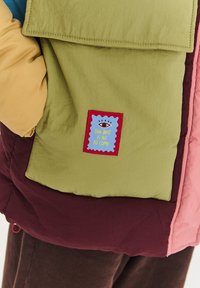 Vícebarevná péřová bunda s zeleným panelem a modrou nášivkou. Nášivka obsahuje grafiku oka a text "THE BEST IS YET TO COME."