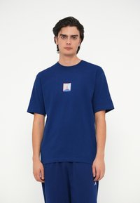 Tumši zils kokvilnas T-krekls ar centrālo logotipu sarkanā un gaiši zilā krāsā. Īsas piedurknes un brīvs piegriezums. Gluda tekstūra.