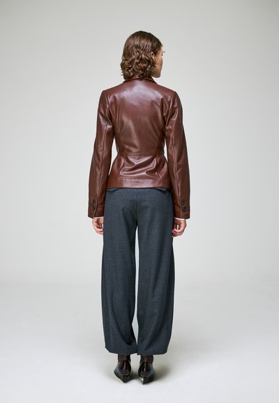 Femme portant une veste en cuir marron cintrée, un pantalon large gris et des chaussures noires pointues, debout face à un fond uni gris clair.