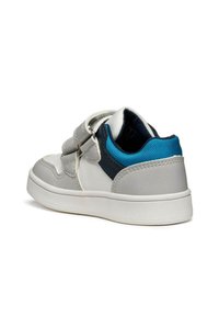 Zapatilla deportiva gris y blanca con acentos azules, que presenta una correa de velcro y punta redonda. Material superior liso y talón texturizado que aportan contraste.