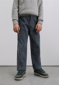 Gray corduroy pants with a textured surface, featuring an elastic waist and wide legs. Paired with dark green sneakers with Velcro straps.