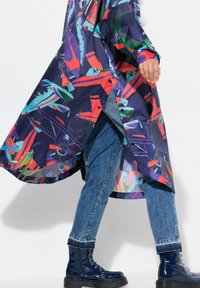 Impermeabile lungo e colorato con motivi astratti in blu navy, rosso, teal e verde. Presenta un orlo asimmetrico ed è abbinato a jeans blu e stivali.