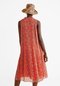 Robe florale rouge avec un design plissé et un col montant, fabriquée en tissu léger. Portée avec un bob à motifs.