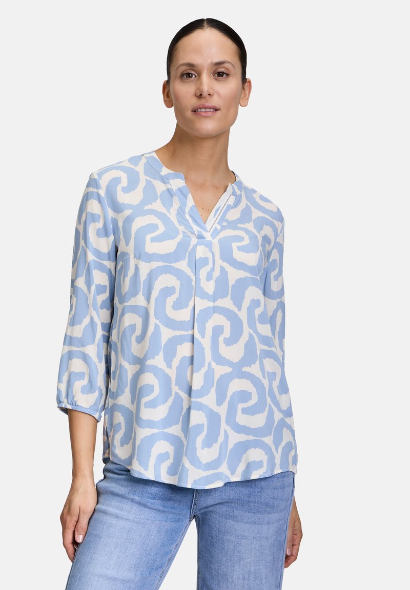 Blusa azzurro chiaro con motivo a spirale bianco, scollatura a V e maniche a tre quarti, realizzata in un tessuto morbido. Abbinata a jeans blu.