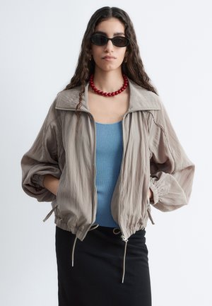 Femme aux longs cheveux bouclés portant des lunettes de soleil noires, un collier de perles rouge, une veste beige zippée, un haut bleu et une jupe noire, mains dans les poches.