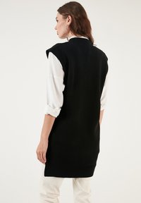 Black knit sleeveless sweater with ribbed hem, worn over a white shirt with rolled sleeves. Smooth texture with relaxed fit and minimalistic design.