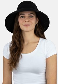 Schwarzer breitkrempiger Hut aus strukturiertem Material mit einem einfarbigen Band. Das Modell trägt ein weißes, eng anliegendes T-Shirt mit kurzen Ärmeln.