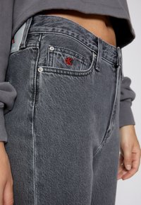 Jeans de mezclilla gris con una textura ligeramente desgastada, que presentan un logotipo bordado rojo en el bolsillo delantero y detalles en hardware plateado.