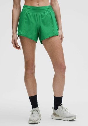 Persona con pantalones cortos deportivos de color verde brillante, calcetines negros y zapatillas blancas de correr, de pie sobre un fondo claro y simple.