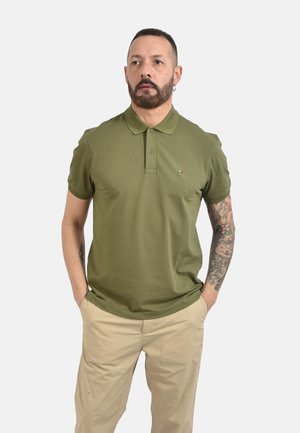 Uomo con barba e braccio destro tatuato indossa una polo verde oliva e pantaloni beige, in piedi con le mani in tasca su uno sfondo uniforme.