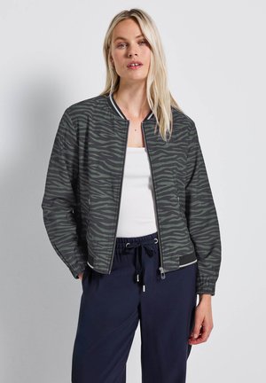 Zebra-patterned bomber jacket in dark green featuring a zipper front, ribbed collar, and elastic cuffs. Paired with navy pants.