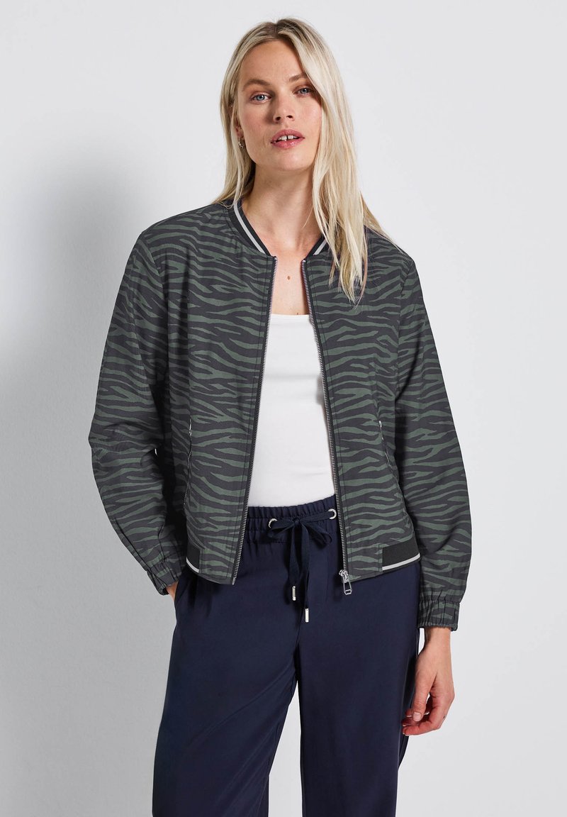 Veste bomber à motif zèbre dans un vert foncé, dotée d'une fermeture éclair à l'avant, d'un col côtelé et de poignets élastiques. Associée à un pantalon bleu marine.
