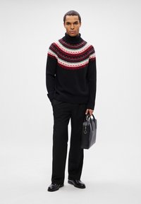 Homme vêtue d'un pull à col roulé noir, rouge et blanc, pantalon noir, chaussures noires, tenant une mallette en cuir noir devant un fond uni.