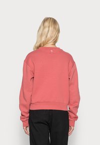 Blondýna v volně střižené korálové mikině a černých kalhotách, pohled zezadu na jednotném světlém pozadí.