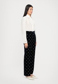 Bílá vyšívaná blůza s límečkem v kombinaci s námořnickými sametovými kalhotami s květinovou výšivkou a úzkým střihem. Modelka má na sobě béžové lodičky.