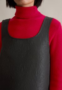 Μαύρο υφασμάτινο φόρεμα με στρογγυλή λαιμόκοψη, το οποίο είναι στρωμένο πάνω από ένα ριμπά τζάκετ σε κόκκινο χρώμα. Το φόρεμα έχει μία διακριτική ανάγλυφη υφή σε όλη του την επιφάνεια.