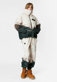 Chaqueta acolchada con cuello alto, con un estampado de paisaje montañoso en beige y verde oscuro. Combinada con pantalones joggers crema y botas marrones.