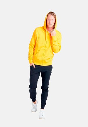 Mann trägt einen gelben Kapuzenpullover mit Reißverschluss, schwarze Jogginghose und weiße Sneakers, steht mit einer Hand in der Tasche.