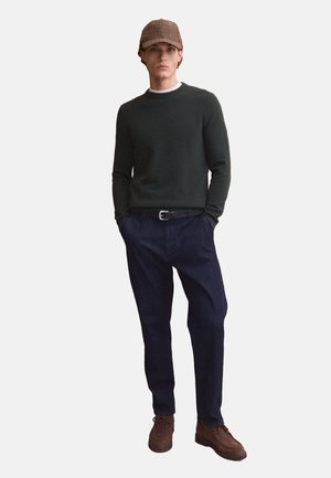 Young man wearing a dark green sweater, navy blue pants, brown shoes, and a brown checkered flat cap, standing with hands in pockets.