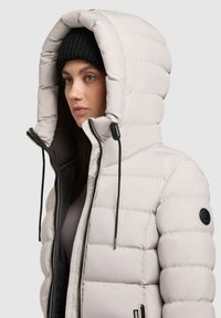 Giubbotto imbottito grigio chiaro con cappuccio e dettagli neri, dotato di chiusura con zip e finitura testurizzata. Include lacci regolabili.