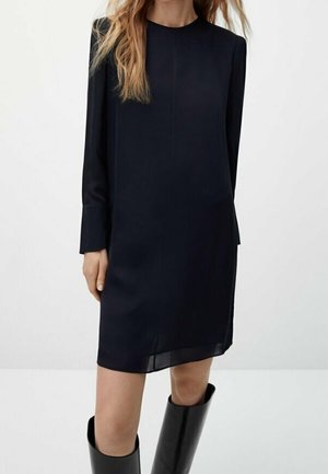 Kobieta ubrana w czarną, długą sukienkę o długości do kolan, w połączeniu z czarnymi kozakami za kolano, stojąca na tle jasnego, jednolitego tła.