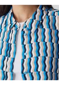 Koszula zapinana na guziki w zygzakowy wzór w odcieniach niebieskiego, beżowego i białego. Posiada kołnierzyk i jasnoniebieskie guziki. Teksturowana tkanina.