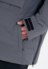 Grå isoleret jakke med en lynlåslomme og velcro-lukning på ærmet. Struktureret stof med vandrette syninger.