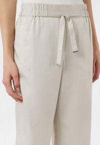 Jasno beżowe spodnie z elastycznym paskiem w talii, z wiązanym sznurkiem i bocznymi kieszeniami. Materiał ma gładką, teksturowaną powierzchnię.