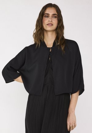 Femme aux cheveux bruns ondulés porte une veste noire courte et ample sur une robe noire plissée, posant devant un fond clair uni.