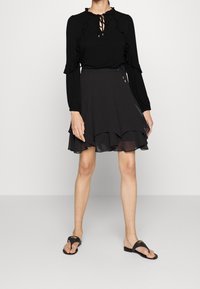 Svart långärmad topp med volanger och nyckelhålsknäppning, ihop med en svart utsvängd kjol med ett lager av genomskinlig nederkant, bärs med svarta flip-flops.