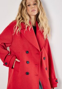 Punainen villasekoitekot joka on kaksirivinen, suurilla kaulusnapeilla ja mustilla nappeilla. Siinä on ylikorostetut hihansuut ja sivutaskut.