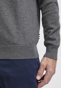 Pulover gri din tricot cu tivuri și manșete ondulate, având o etichetă mică de marcă cu dungi roșii și albastre pe lateral, purtat peste pantaloni bleumarin.