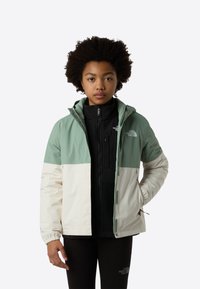 Veste à blocs de couleurs en vert clair et crème, avec une couche intérieure noire. Comprend une capuche, des poignets élastiques et des poches zippées.