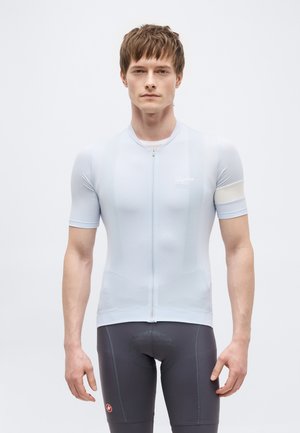 Ung mann iført en lyseblå sykkeltrøye med korte ermer og mørkegrå polstrede sykkelshorts, stående mot en ensfarget bakgrunn.