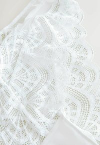 Balta mežģīņu bralete ar sarežģītām ziedu rakstām, caurspīdīgu tekstūru un gludām auduma detaļām. Izliekti malām piešķir dizainam maigumu.