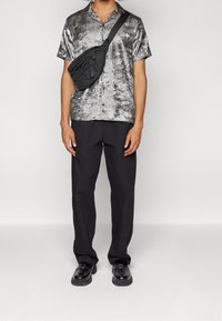 Camicia metallizzata argento a maniche corte con colletto, abbinata a pantaloni neri larghi e scarpe nere slip-on, completata da una borsa a tracolla nera.