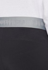 Melni leggins ar plašu, gaiši pelēku jostu, uz kuras ir uzraksts "THE NORTH FACE". Audums izskatās gluds ar nelielu spīdumu.