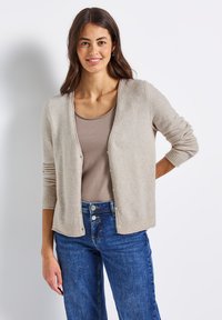 Kvinna med långt brunt hår som bär en beige cardigan över en taupe-top och blå jeans, ståendes med en hand på höften mot en vit bakgrund.
