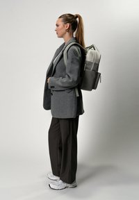 Blazer grigio in lana, pantaloni marroni larghi e sneakers bianche. Uno zaino leggero grigio chiaro con più scomparti indossato sulla schiena.