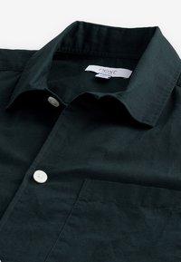 Camisa verde oscura con un collar estructurado, botones blancos y un bolsillo en el pecho. Fabricada con una tela suave y texturizada. Etiqueta visible dentro del collar.