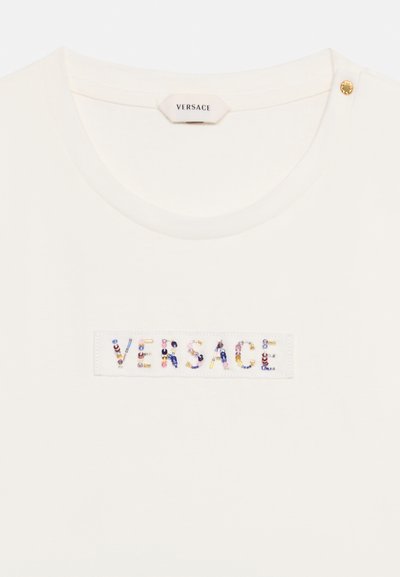 Λευκό τοπ Versace με πολύχρωμο κεντημένο όνομα επωνυμίας στο στήθος και χρυσή λεπτομέρεια με κουμπί κοντά στο ντεκολτέ.