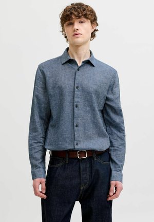 Jonge man met krullend haar, gekleed in een blauw overhemd met knopen en lange mouwen, donkere spijkerbroek en een bruine riem, tegen een effen achtergrond.