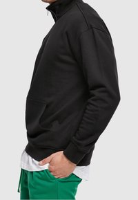 Sudadera negra con cremallera y alto cuello, con bolsillos laterales y puños acanalados, superpuesta sobre una camiseta blanca y combinada con pantalones de chándal verdes.