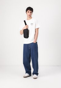 Schwarze Schultertasche, weißes T-Shirt mit Logo, lässige blaue Jeans und helle Sneakers. Hintergrund ist rein weiß.