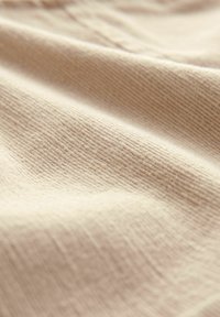Beige teksturoitu kangas, jossa on pehmeä, poimuttunut pinta, joka esittää hienovaraisia viivoja ja aaltoilevia taitoksia, mikä osoittaa joustavuutta ja pehmeyttä.