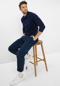 Camicia a maniche lunghe blu navy abbinata a pantaloni blu scuro; modello seduto su uno sgabello di legno. Sneakers bianche e calzini color crema completano l'outfit.
