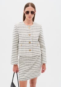 Bílá a námořnická pruhovaná bunda se zlatými knoflíky, spojená s ladícími sukní. Model má na sobě sluneční brýle a drží černou kabelku.